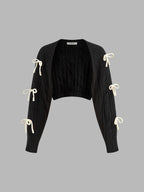A black cropped cable knit cardigan with open front and three white ribbon bows per sleeve.