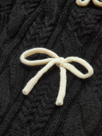 A black cable knit cardigan with intricate patterns and a white bow-knot accent on top.